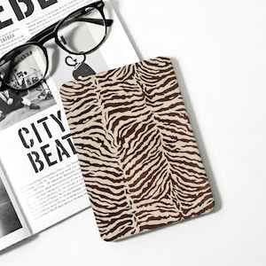 May include: A rectangular e-reader cover featuring a brown and cream tiger stripe pattern. The cover is positioned next to an open magazine with black and white text and a pair of black-rimmed spectacles resting on the top left corner of the page.