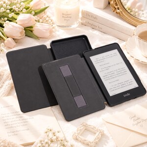 Puede incluir: Una funda negra para lector electr&oacute;nico Kindle con tapa plegable y correa de mano integrada en la parte posterior. La funda se muestra abierta, revelando la estructura interior y la pantalla del dispositivo con texto en franc&eacute;s.