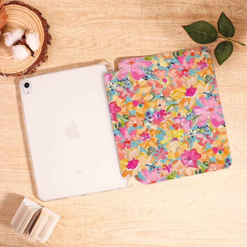 May include: A white iPad with a clear case next to a floral patterned case. The floral pattern features pink, yellow, and orange flowers on a light blue background. The iPad is on a light brown wooden surface.