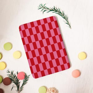 Könnte beinhalten: Ein rechteckiger Gegenstand mit einem rosa-roten Schachbrettmuster. Der Gegenstand ist von bunten, runden Objekten und Zweigen auf einer weißen Oberfläche umgeben. Das Muster ist ein sich wiederholendes Gitter aus abwechselnd rosa und roten Rechtecken.