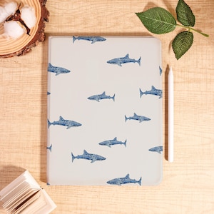 May include: A light gray tablet case featuring a repeating pattern of blue shark illustrations. A white stylus rests beside the case on a wooden surface. The case is rectangular and appears to be made of a smooth material.