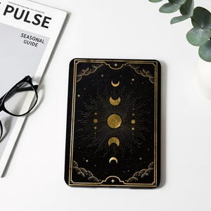 Può includere: Una custodia rettangolare per e-reader con sfondo nero e design celeste dorato. La grafica mostra una luna piena centrale circondata da varie fasi lunari, raggi solari radianti, piccole stelle e bordi decorativi a forma di nuvola.
