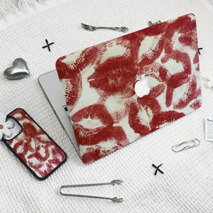 May include: A laptop and phone case with a red lipstick kiss print on a white background. The laptop has a white Apple logo. A silver heart, fork, and other accessories are scattered around the items.