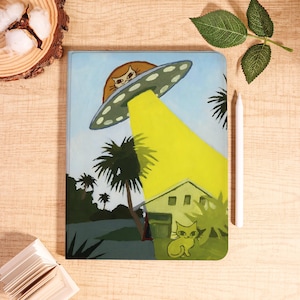 Op de afbeelding: Een kleurrijk notitieboek met een grillige scène van een kat die door een UFO wordt ontvoerd. De illustratie bevat palmbomen, een huis en een gele lichtstraal. Een witte stylus ligt ernaast.