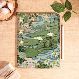Op de afbeelding: Een decoratief notitieboek met een kikker op een waterlelie, omringd door waterlelies en madeliefjes. De cover heeft een vintage esthetiek met de woorden "healing happens". Een witte stylus en lichtslingers met stervormige ornamenten zijn ook aanwezig.
