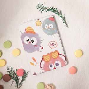 May include: White rectangular phone case featuring cartoon characters with expressive eyes and accessories. The case has a light purple, pink, and blue color scheme with the text "Hi~".