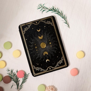 May include: A black rectangular tarot card with a gold celestial design featuring phases of the moon, stars, and sunburst. The card is framed with a gold border and decorative clouds. The background is white with colorful macarons and greenery.