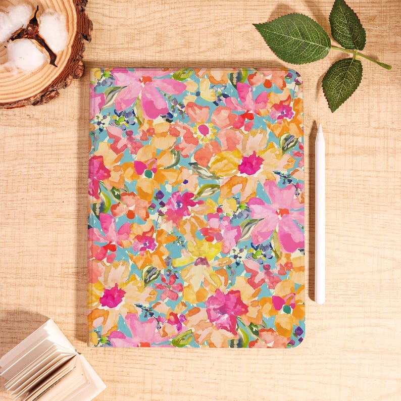 May include: A floral-patterned tablet case with a vibrant design featuring pink, orange, and yellow flowers on a light blue background. A white stylus rests beside the case. The case is rectangular and likely designed for an iPad or similar device.