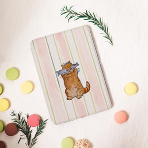 May include: A rectangular case with a striped pattern in pink, white, and green. A cartoon orange tabby cat with two fish in its mouth is centered on the case. The case is surrounded by colorful macarons and greenery.