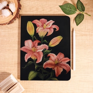Könnte beinhalten: Ein rechteckiges Notizbuch mit schwarzem Einband, das mit einer gemalten Illustration von rosa Lilien, grünen Stielen und gelben Knospen verziert ist. Das Notizbuch liegt vertikal mit einem weißen Eingabestift rechts daneben auf hellem Holz.