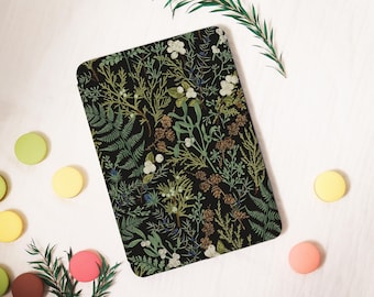 Puede incluir: Un cuaderno rectangular negro con un diseño botánico de helechos, bayas y follaje en tonos verdes, blancos y marrones. El cuaderno está rodeado de macarons coloridos y ramitas verdes sobre una superficie blanca.