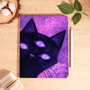 May include: A notebook with a black cat illustration, featuring three eyes and a purple floral design. A white stylus is placed next to the notebook. The cat's eyes are light pink with a circular design. The notebook is on a wooden surface.