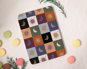 May include: A rectangular mat featuring a celestial design with suns and moons in a chequered pattern. The colours include orange, purple, and blue. The mat is surrounded by colourful macaroons and greenery.