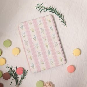 Peut inclure: Une housse de tablette rectangulaire à motifs floraux, avec un design à rayures roses et blanches. La housse présente de délicates fleurs bleues, jaunes et blanches. L'image est agrémentée de macarons colorés et de verdure sur fond blanc.