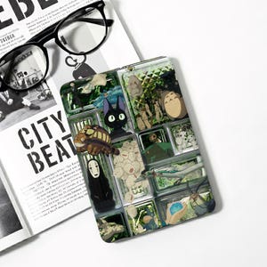 May include: A rectangular e-reader cover featuring a collage of various animated characters, including a black cat, a large grey creature, a white masked figure, and small white spirits, all arranged in a grid-like pattern with green accents.