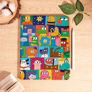May include: A colorful notebook featuring a grid of cartoon characters in various shapes and colors. The cover design includes a variety of whimsical faces and figures, with a white stylus resting beside it. The notebook is on a wooden surface.