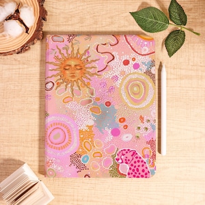 May include: A pink and peach-colored journal with an abstract design featuring a sun, dots, and a pink cheetah. A white stylus rests beside the journal. The journal is on a wooden surface.