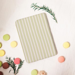 Op de afbeelding: Een rechthoekige hoes met een lichtgroen en wit gestreept patroon, mogelijk voor een tablet of e-reader, is omgeven door kleurrijke macarons en takjes groen op een wit oppervlak. De macarons zijn in tinten groen, geel, roze en bruin.