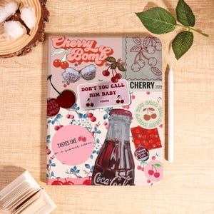 May include: A decorative notebook with a collage of cherry-themed graphics, including a Coca-Cola bottle, cherries, and the text "Cherry Bomb." The notebook has a pink and red color scheme and a white stylus.