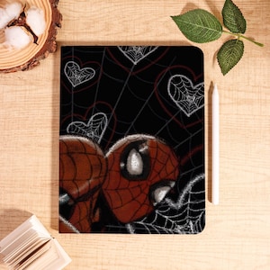 May include: A black notebook featuring a Spiderman design with a web and heart pattern. The cover showcases the character's mask in red and black, with white web details and heart-shaped webs. A white stylus rests beside the notebook.