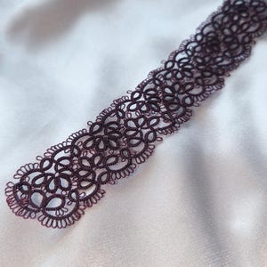 May include: A dark burgundy lace choker necklace with an intricate floral design. The delicate, openwork pattern is displayed against a white satin background, highlighting the detailed craftsmanship of the jewellery.