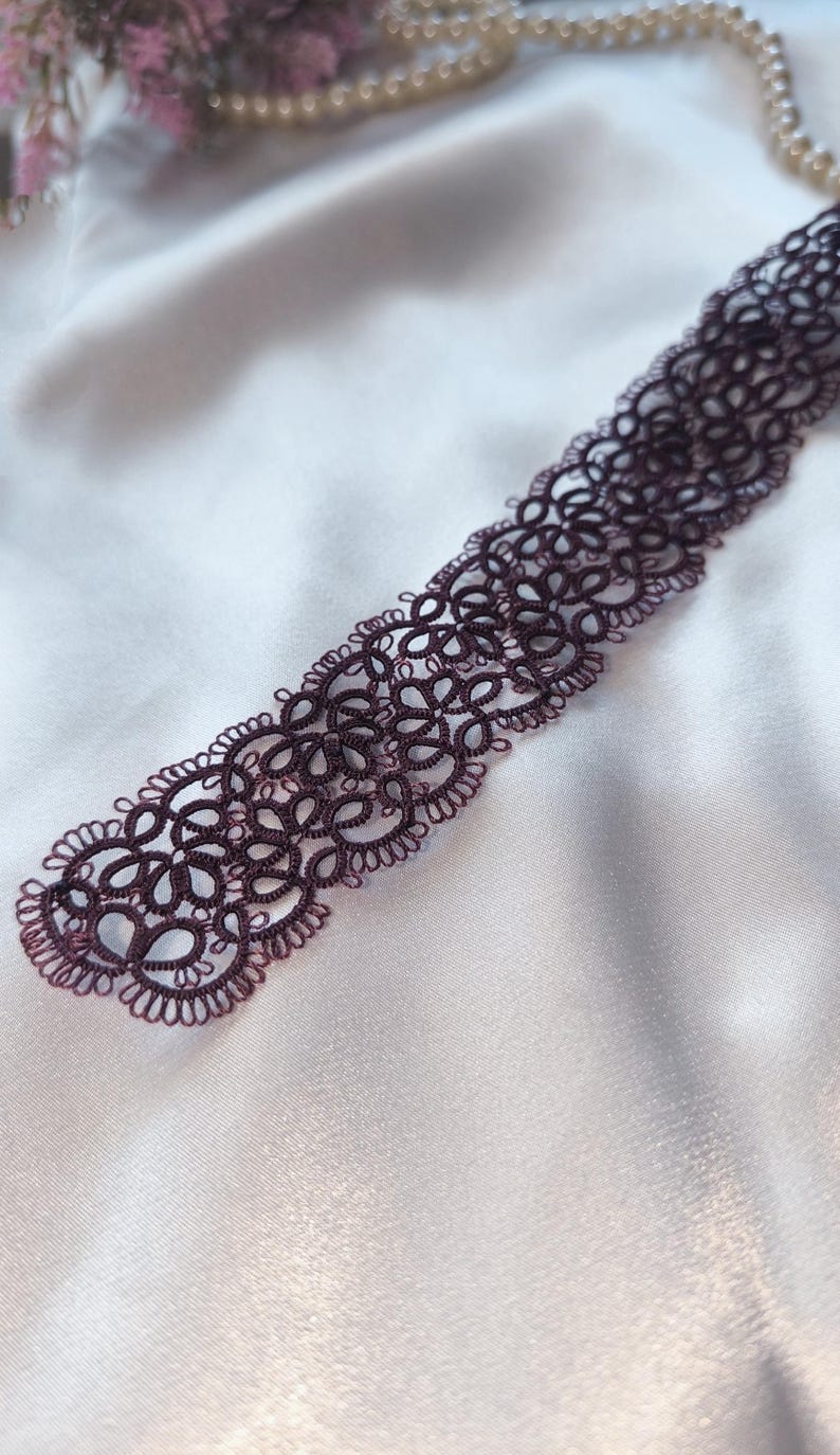 May include: A dark burgundy lace choker necklace with an intricate floral design. The delicate, openwork pattern is displayed against a white satin background, highlighting the detailed craftsmanship of the jewellery.