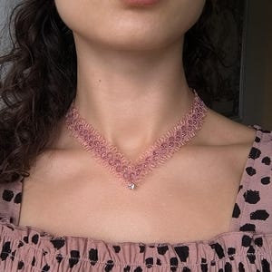Puede incluir: Un collar gargantilla de encaje rosa con un diseño delicado y pequeños detalles de cuentas. El collar tiene forma de V y un pequeño colgante. La modelo lleva un top rosa con lunares negros y un pendiente azul.