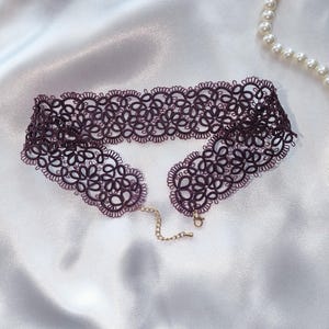 May include: A deep purple lace choker necklace with a floral design. It features a gold-coloured chain and clasp. The background is white satin fabric, with a pearl necklace and pink flowers.