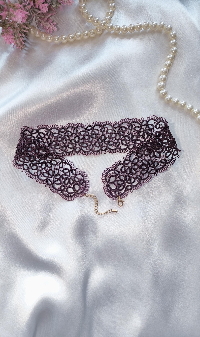 May include: A deep purple lace choker necklace with a floral design. It features a gold-coloured chain and clasp. The background is white satin fabric, with a pearl necklace and pink flowers.