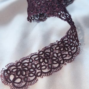 May include: A length of deep purple lace ribbon with an intricate, openwork design. The ribbon is curled and lies on a white satin surface, with a blurred background of pearls and flowers.