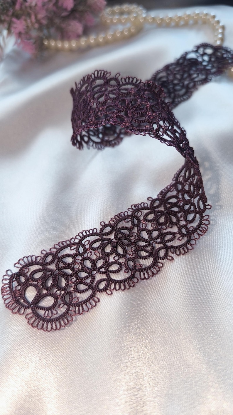 May include: A length of deep purple lace ribbon with an intricate, openwork design. The ribbon is curled and lies on a white satin surface, with a blurred background of pearls and flowers.