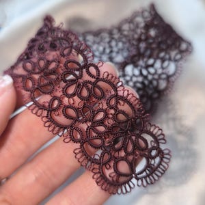 May include: Close-up of a piece of dark burgundy lace with a floral design. The lace is held in a hand, showcasing its intricate details and delicate texture. The background is blurred, highlighting the lace.