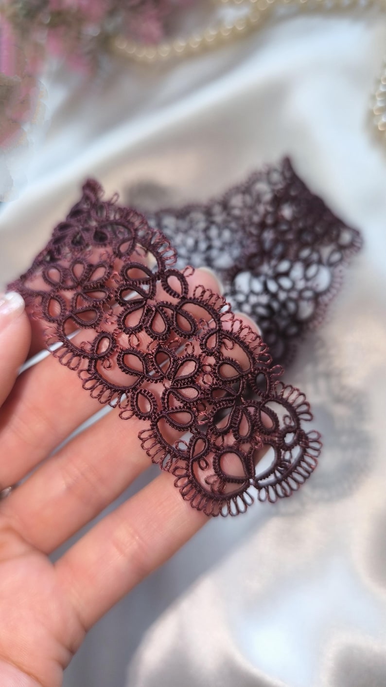 May include: Close-up of a piece of dark burgundy lace with a floral design. The lace is held in a hand, showcasing its intricate details and delicate texture. The background is blurred, highlighting the lace.