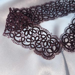 May include: A dark burgundy lace choker necklace with an intricate floral design. The delicate openwork pattern is displayed against a white satin background, with a pearl necklace and pink flowers in the background.