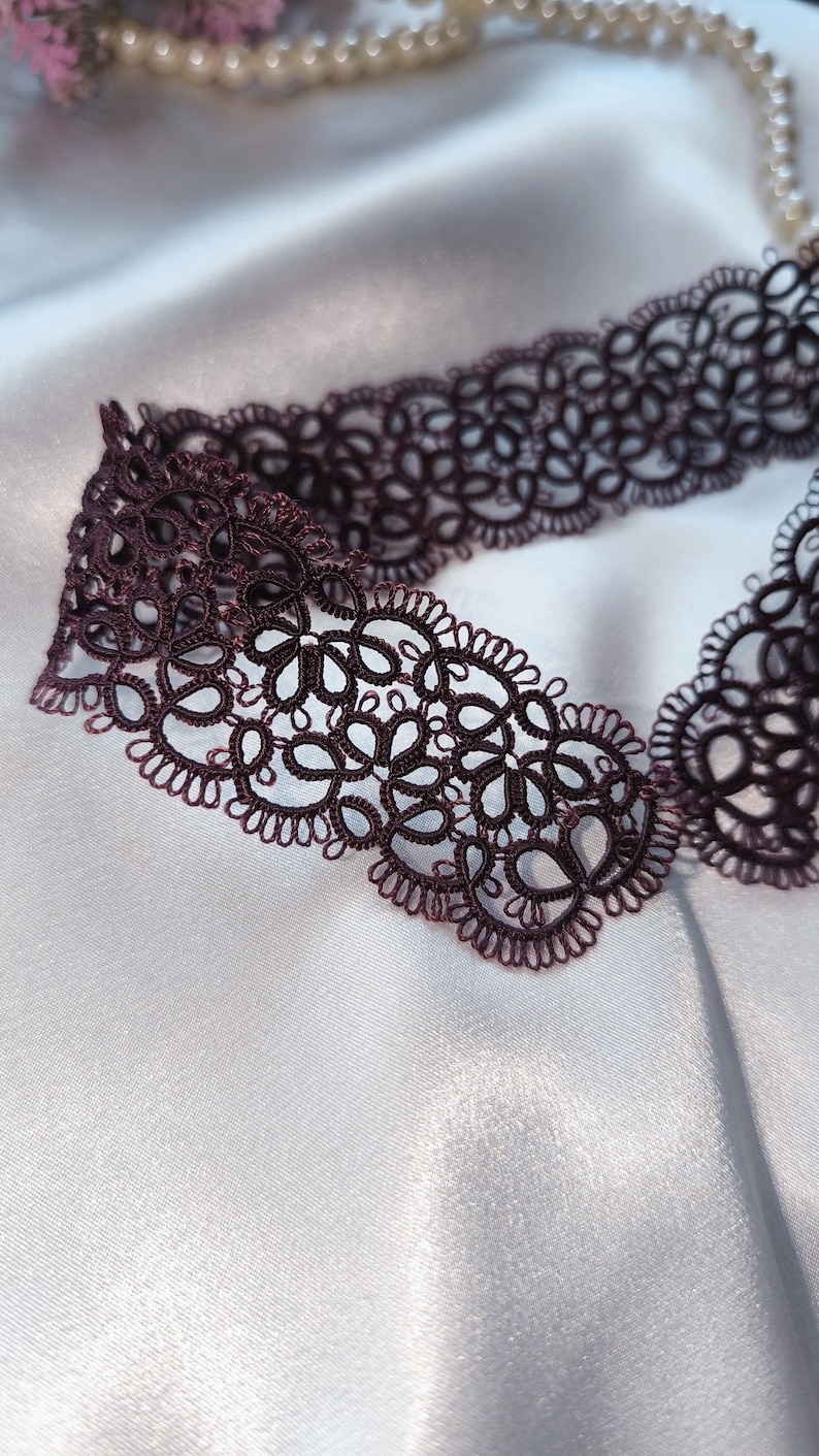 May include: A dark burgundy lace choker necklace with an intricate floral design. The delicate openwork pattern is displayed against a white satin background, with a pearl necklace and pink flowers in the background.