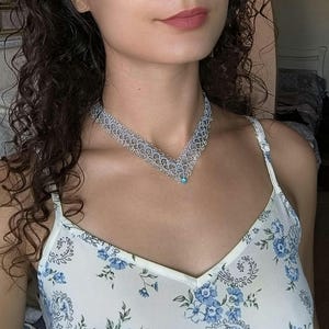 May include: A silver-colored necklace with a V-shaped design. The necklace has a delicate, lacy pattern and a small, round, blue gemstone at the center. The person is wearing a white camisole with a blue floral pattern.