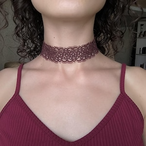 May include: A dark red lace choker necklace featuring a floral pattern. The necklace is worn around the neck, paired with a matching burgundy vest top. The image is a close-up of the neck and upper chest.