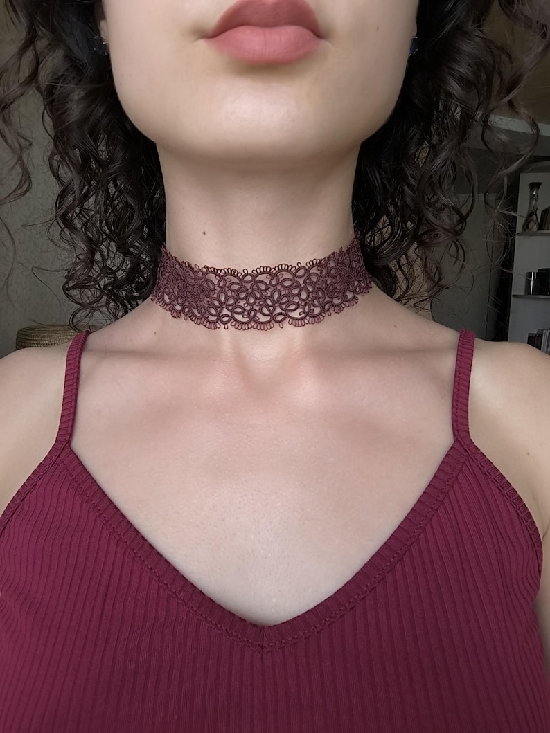 May include: A dark red lace choker necklace featuring a floral pattern. The necklace is worn around the neck, paired with a matching burgundy vest top. The image is a close-up of the neck and upper chest.