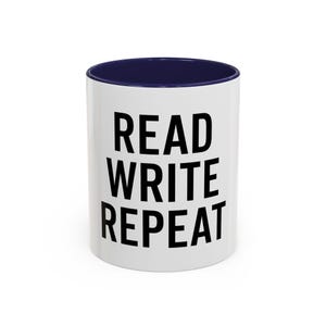 May include: A white ceramic mug with a dark blue interior and rim. The exterior features the black text READ WRITE REPEAT printed in a bold, sans-serif font. The mug has a cylindrical shape and a smooth, glossy finish on the outer surface.