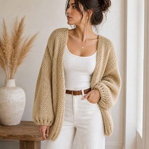 Può includere: Un cardigan beige a maglia grossa con maniche lunghe e voluminose. Il maglione aperto è indossato sopra una canotta bianca e abbinato a pantaloni bianchi e una cintura marrone. L'outfit è completato da una collana d'oro.