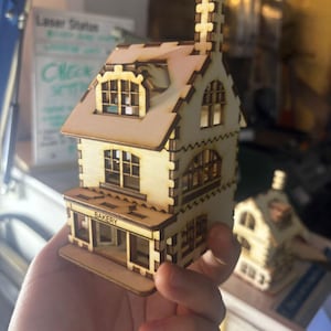 May include: A detailed miniature wooden bakery model, laser-cut with precision. The two-storey building has multiple windows, a chimney, and a sign reading "BAKERY". The model is held in a hand, highlighting its size and craftsmanship.