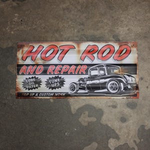 May include: A rectangular metal sign with a distressed finish, featuring the words "HOT ROD AND REPAIR" in red and white. Below, a black and white illustration of a classic car is displayed. Additional text includes "LUBE OIL FILTER" and "TUNE UPS".