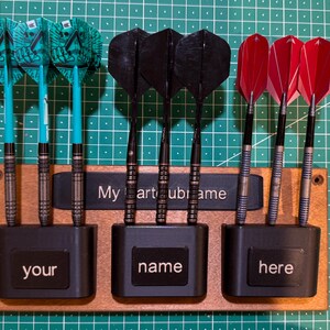 May include: A wooden dart holder with three sections, each holding three darts. The darts have teal, black, and red flights. The holder has the text "your", "name", and "here". The text "My art ubi name" is above the sections.