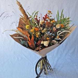 Puede incluir: Un ramo de flores secas en tonos marrones, naranjas y amarillos, envuelto en papel marrón y atado con una cinta negra.
