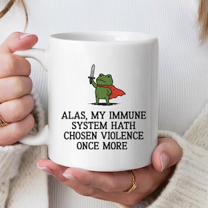 May include: White ceramic mug featuring a cartoon frog with a cape and sword. The text on the mug reads: "ALAS, MY IMMUNE SYSTEM HATH CHOSEN VIOLENCE ONCE MORE". The frog is green, the cape is red, and the text is black.