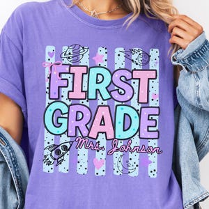 Puede incluir: Una camiseta violeta de manga corta con un estampado gráfico que incluye el texto FIRST GRADE y Mrs. Johnson. El diseño presenta rayas verticales, un cohete, planetas, estrellas y corazones en tonos azul claro, rosa y violeta.
