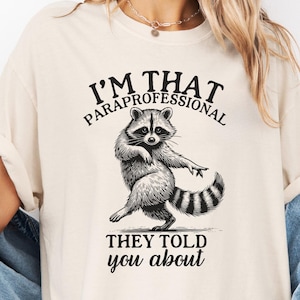 May include: Ivory-coloured t-shirt featuring a dancing raccoon graphic and the text "I'M THAT PARAPROFESSIONAL THEY TOLD you about". The shirt is made of Comfort Colors material.