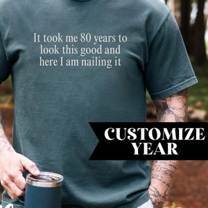 May include: A dark teal short-sleeve t-shirt featuring the white printed text: It took me 80 years to look this good and here I am nailing it. The shirt has a crew neckline and a standard fit, showcasing a solid color design throughout the fabric.
