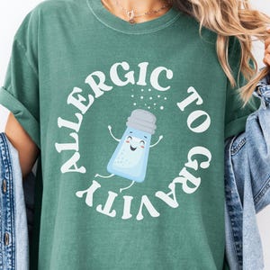 以下が含まれることがあります： 白い文字で「重力アレルギー」と書かれた薄い緑色のTシャツ。テキストの中央に笑顔の塩入れの漫画が描かれている。