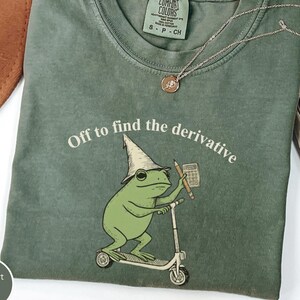 May include: A moss green t-shirt featuring a graphic of a frog wearing a pointed hat, riding a scooter, and holding a pencil and calculator. The text above the frog reads, Off to find the derivative. The shirt has a Comfort Colors brand label.